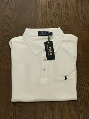 Vit pikétröja från Polo Ralph Lauren - Klassisk vit pikétröja från Polo Ralph Lauren med broderad marinblå logga på bröstet. Tröjan har krage, knappar framtill och är tillverkad i ett strukturerat bomullstyg. Perfekt för en clean och stilren look.