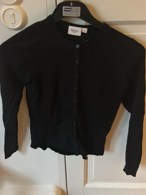 Svart cardigan från Saint - Klassisk svart cardigan från Saint med rund halsringning och knäppning framtill. Långärmad modell i mjukt och tunt material som passar perfekt till lager på lager. Enkel och stilren design som funkar till det mesta. En knappt har gått sönder men funkar normalt