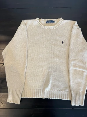 Beige stickad tröja Polo Ralph Lauren - Säljer en beige stickad tröja från Polo Ralph Lauren med klassisk rund hals och diskret broderad logga på bröstet. Tröjan har ribbade muddar vid ärmslut och nederkant, perfekt för lager på lager under höst och vinter.