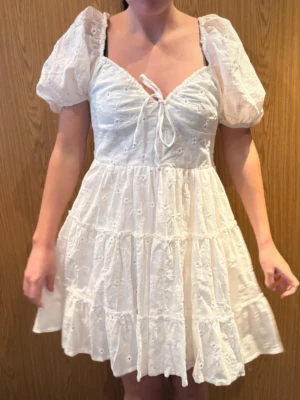 Vit broderad kortklänning Chiquelle XS Studentklänning - Superfin vit kortklänning från Chiquelle i storlek XS. Klänningen har puffärmar, v-ringning med knyt och broderade blommor över hela tyget. Den är sydd i flera volanger och har en romantisk känsla, perfekt för sommarens alla tillfällen. Perfekta studentklänningen! 