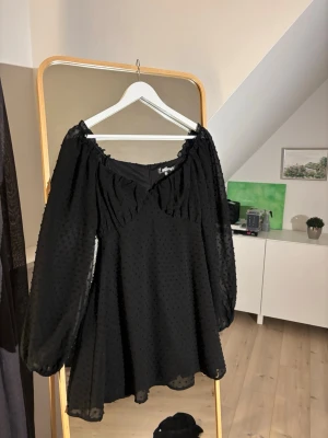 Svart prickig klänning med volang - Supersöt svart klänning med transparenta, långärmade ärmar och diskret prickigt mönster. Klänningen har en djup v-ringning med volangdetalj och luftig passform. Perfekt för dig som gillar romantisk och trendig stil.