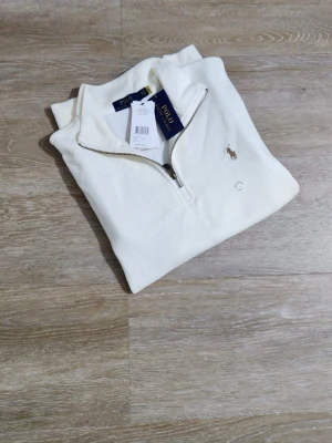 Vit half zip tröja från Polo Ralph Lauren - Säljer en stilren vit tröja från Polo Ralph Lauren med klassisk half zip-dragkedja och broderad logga på bröstet. Tröjan är i mjuk bomull och har långa ärmar samt ribbade muddar. Perfekt för dig som gillar clean och tidlös stil.