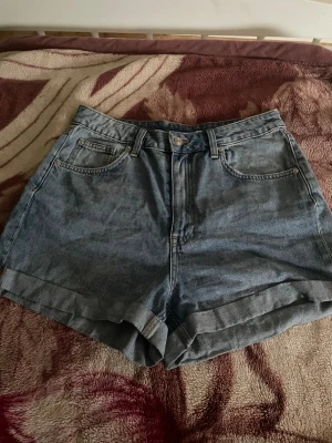 Högmidjade denimshorts i blått - Snygga högmidjade denimshorts i klassisk blå färg med uppvikta benslut. Shortsen har fem fickor och stängs med knapp och dragkedja. Perfekta för varma dagar och enkla att matcha med olika toppar.