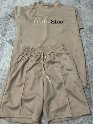 Beige set med texten Don't Stop - Snyggt beige set med t-shirt och shorts i mesh-liknande material. T-shirten har trycket 'Don't Stop' framtill och rund hals. Shortsen har resår och dragsko i midjan för skön passform. Perfekt för en avslappnad och sportig stil.