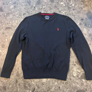 Mörkblå tröja från Polo Ralph Lauren - Mörkblå tröja från Polo Ralph Lauren med klassisk rund hals och röd broderad logga på bröstet. Tröjan har långa ärmar och ribbade muddar vid ärmslut och nederkant. Storlek M (10-12 år).
