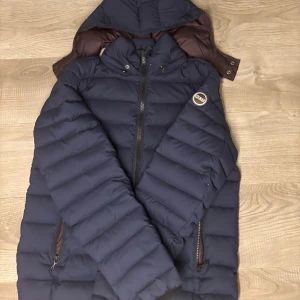 Mörkblå dunjacka från Colmar - Säljer en snygg mörkblå dunjacka från Colmar med avtagbar huva och dragkedja framtill. Jackan har två fickor med dragkedja och ett brunt innerfoder. Perfekt för kalla dagar och riktigt skön att bära.