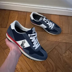 Blå och vita sneakers från Polo Ralph Lauren - Snygga sneakers från Polo Ralph Lauren i marinblått, grått och vitt med röd detalj på sulan. Skorna har klassisk snörning, vit logga med polospelare på sidan och en platt sula. Ovandelen är i mocka och textil för en sportig och retro vibe.
