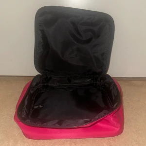 Rosa necessär från Victoria's Secret - 25 cm på bredden. 20 cm på längden och 8 cm på djupen. Snygg och praktisk necessär från Victoria's Secret i stark rosa färg med glansig satinfinish. Väskan har en rektangulär form, svart insida och dragkedja runt om. Perfekt för smink eller småprylar när du är på språng. 