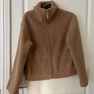 Beige teddyjacka med dragkedja - Mysig beige teddyjacka från HM med hög krage och hel dragkedja framtill. Jackan har två stora fickor och är tillverkad i mjuk fleece som håller dig varm under kyliga dagar. Perfekt för lager-på-lager och chill vardagsstil.