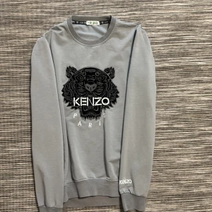 Grå Kenzo sweatshirt med tigerbrodyr - Säljer en grå sweatshirt från Kenzo med ikonisk svart tigerbrodyr och vit Kenzo Paris-text på bröstet. Tröjan har rund hals, långa ärmar och ribbade muddar. Tillverkad i mjuk bomull med lite stretch för extra komfort.