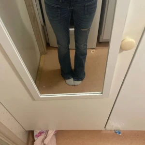 Blå bootcut jeans med slitningar - Säljer ett par blå jeans med bootcut-modell och snygga slitningar vid knäna. Jeansen har låg midja och klassiska fickor fram och bak. Perfekta för dig som gillar en avslappnad och trendig look.