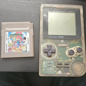 Nintendo Game Boy Pocket Transparent + Super Mario Land 2 - Nintendo Game Boy Pocket i transparent utförande med Super Mario Land 2: 6 Golden Coins. Konsolen visar tecken på användning och viss missfärgning. Testad och funkar. Batterilucka saknas. Perfekt för retrospelare och samlare! OBS HÄMTAS PÅ PLATS! 