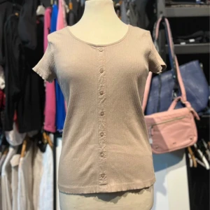 Beige ribbad topp med knappar - Snygg beige ribbad topp från Black story med korta ärmar och rund halsringning. Toppen har knappar framtill som en fin detalj och är tillverkad i mjuk bomull. Storlek S/M