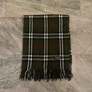 Grön rutig ullhalsduk från Burberry - Stilren halsduk från Burberry i mörkgrönt med klassiskt rutigt mönster i vitt, svart och rött. Gjord i mjuk ull och har snygga fransar i ändarna. Perfekt accessoar för att lyfta din outfit med en tidlös touch.