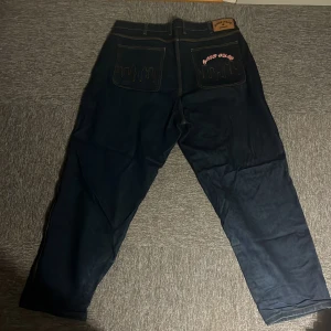 Cash Only mörkblå jeans med broderi - Mörkblå jeans från Cash Only med coolt broderi av stadssilhuett på bakfickorna och röd 'Cash Only'-text. Framficka har ett broderat dollartecken. Klassisk femficksmodell med stora ben och baggy passform. Perfekt för dig som gillar streetwear och unika detaljer.