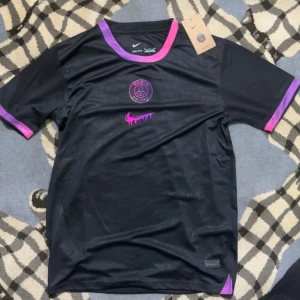  PSG  fotbollströja - Snygg svart fotbollströja från Nike i samarbete med Paris Saint-Germain. Tröjan har lila och rosa detaljer vid hals och ärmslut, samt PSG-logga och Nike Swoosh i rosa på bröstet. Tillverkad i lätt och ventilerande material, perfekt för träning eller match.