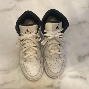 Nike Air Jordan 1 Mid vit/svart - Säljer ett par Nike Air Jordan 1 Mid sneakers i vit och svart. Klassisk design med snörning, perforerad tå och svart Swoosh på sidan. Skorna har en rund tå och är tillverkade i skinn med svart foder och detaljer. Perfekt för dig som gillar streetstyle och ikoniska sneakers.