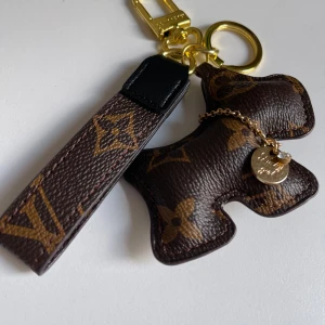  Nyckelring med hund och väska accessoarer  - Lyxig nyckelring från  Vuitton med klassiskt monogrammönster i brunt och guld. Nyckelringen har en söt hundformad detalj och guldfärgade metalldelar, samt en liten rund berlock. Perfekt som accessoar till väskan eller nycklarna.