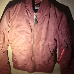 Mörkrosa bomberjacka Alpha Industries S - Säljer en snygg mörkrosa bomberjacka från Alpha Industries i storlek S. Jackan har klassisk ribbad krage, dragkedja framtill, fickor med tryckknappar och en ikonisk röd tag på ärmen. Materialet är glansig polyester och jackan är quiltad för extra värme.