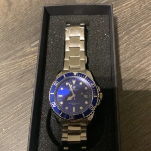 Blå och silverfärgad klocka  - Stilren klocka från Kompass Stockholm med blå urtavla, silverfärgad boett och länkarmband i metall. Klockan har tydliga index, datumvisning och vridbar ring. Kommer i originalask med tillbehör och garanti. Endast provad en gång sen har den bara legat. Nypris 1200kr mitt pris 700kr