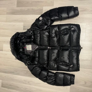 Moncler maya  - Snygg svart dunjacka från Moncler med glansig finish, huva och flera dragkedjeförsedda fickor. Jackan har klassisk Moncler-logga på ärmen och coolt serietryck på insidan. Perfekt för kalla dagar när du vill hålla stilen. Skicken är bra. Pris kan diskuteras.