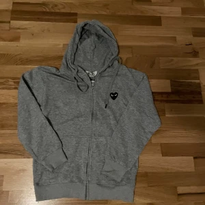 Grå hoodie från Comme des Garçons Play - Grå hoodie med dragkedja från Comme des Garçons Play. Tröjan har huva med snörning, svart hjärta med ögon på bröstet och stort svart hjärta med ögon på ryggen. Tillverkad i mjuk bomull och har långa ärmar samt ribbade muddar.