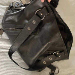 Svart axelväska i skinn med ringdetaljer - Cool svart axelväska i skinn med flera silvriga ringdetaljer. Vintage super snygg. Den har både korta band och ett långt axelband så man kan ha den över kroppen. Används tyvärr inte. Trasig på ena sidan då den har tappat en knapp på ena sidan (ser ni bild 3 o 4). 