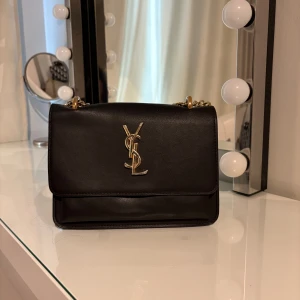 Svart YSL axelväska med guldkedja - Snygg svart axelväska från YSL i slätt skinn med guldfärgad kedja och ikonisk YSL-logga framtill. Väskan har flera fack invändigt med vinrött innerfoder och magnetlås. Perfekt storlek för det viktigaste och ger en lyxig vibe till din outfit.