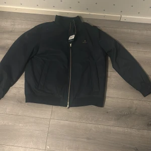 Mörkblå bomberjacka från Gant XL - Snygg mörkblå bomberjacka från Gant i modellen The Hampshire Jacket. Jackan har klassisk rak passform, ståkrage, två sidofickor och hel dragkedja framtill. Fodret är i kontrasterande ljusblått och mörkblått. Perfekt för dig som vill ha en stilren och tidlös look.