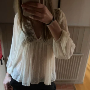 Vit blus med paljettdetaljer - Romantisk vit blus i tunn bomull med diskret prickigt mönster och v-ringning. Blusen har långa, luftiga ärmar och är dekorerad med silvriga paljetter och pärlor i blommönster vid halsringningen. Perfekt för dig som gillar boho-vibes och snygga detaljer.