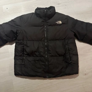 Svart pufferjacka från The North Face - Säljer en svart pufferjacka från The North Face i storlek L. Jackan har hög krage, dragkedja framtill och två sidofickor. Klassisk logga broderad på bröstet och ryggen. Perfekt för kalla dagar med sin vadderade design.