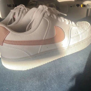 Nike /rosa sneakers - Fräscha Nike sneakers i vitt med ljusrosa swoosh och häl. Klassisk låg modell med snörning, perforerad tå och platt sula. Skorna är tillverkade i skinn och har en rund tå. Perfekt för dig som gillar stilrena och trendiga sneakers.