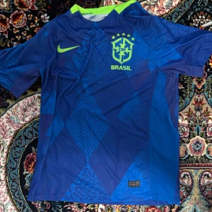 Brasilien fotbollströja - Snygg blå Brasilien fotbollströja från Nike med neongula detaljer och CBF-emblem på bröstet. Tröjan har korta ärmar och ett unikt geometriskt mönster i olika blå nyanser. Perfekt för dig som vill sticka ut på planen eller på läktaren.
