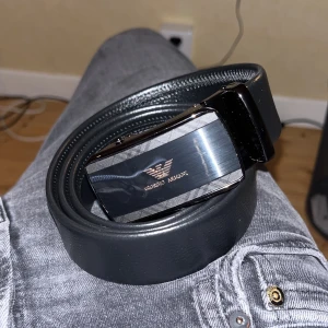 Svart skärp från Giorgio Armani - Snyggt svart bälte från Giorgio Armani med stilren, blank metallspänne och logga framtill. Bältet är gjort i slätt skinn och har en modern, minimalistisk look som passar till jeans eller kostymbyxor. Perfekt accessoar för att lyfta din outfit.