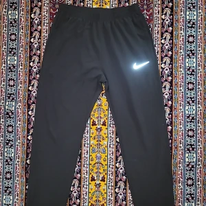 Svarta träningsbyxor från Nike - Svarta träningsbyxor från Nike med reflekterande logga på låret. Byxorna har dragkedjeförsedda fickor och elastisk midja för skön passform. Perfekta för träning eller chill, tillverkade i mjukt och lätt syntetmaterial.