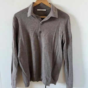 Pikétröja i ull från A Day's March - Grå/brun pikétröja från A Day's March. Tröjan har klassisk krage, knäppning med två knappar och långa ärmar. Storlek M men sitter som S! Hade inte köpt om man brukar ha M i tröjor. 