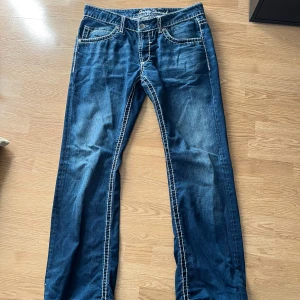 Blåa jeans från Camp David med kontrastsömmar - Snygga blå jeans från Camp David med tydliga vita kontrastsömmar och klassisk femficksdesign. Jeansen har raka ben och coola detaljer på bakfickorna. Perfekta för dig som gillar en avslappnad men stilren look.