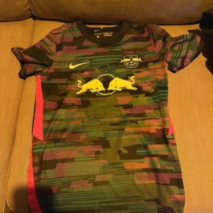 RB Leipzig Nike fotbollströja mönstrad - Säljer en RB Leipzig fotbollströja från Nike med coolt digitalt mönster i grönt, rött och svart. Tröjan har korta ärmar, rund hals och är tillverkad i lätt och ventilerande Dri-FIT material. Stort Red Bull-tryck på bröstet och klubbmärke på vänster sida.