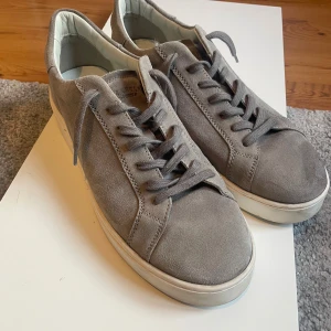 Grå mocka sneakers från Stockholm Design Group - Snygga grå sneakers i mocka från Stockholm Design Group. Skorna har en klassisk låg profil, rund tå och vita platta sulor. De har snörning framtill och en stilren design som passar till det mesta. Perfekta för dig som gillar en clean och modern look.