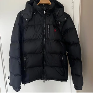 Svart dunjacka Polo Ralph Lauren M - Svart dunjacka från Polo Ralph Lauren med röd logga på bröstet. Jackan har huva, flera dragkedjor och en quiltad design. Perfekt för kalla dagar och har en klassisk, stilren look som funkar till det mesta.