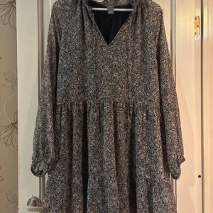 Blommig långärmad klänning från Lindex - Supersöt långärmad klänning från Lindex med blommigt mönster i blått och beige. Klänningen har v-ringning, lös passform och faller snyggt med lätt rynk vid midjan. Perfekt för dig som gillar en avslappnad men ändå stilren look.