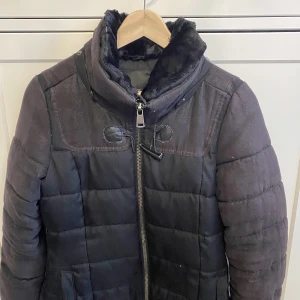 Svart pufferjacka med fuskpäls från Amisu - Säljer en svart pufferjacka från Amisu med mysig fuskpäls i kragen och quiltad design. Jackan har dragkedja framtill, hög krage och två fickor med knappstängning. Perfekt för kalla dagar när du vill hålla dig varm och snygg.