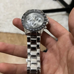 Kronografklocka från BOSS i silver - Stilren kronografklocka från BOSS med rund svart urtavla, tre mindre subdials och tydliga index. Armbandet är i blankt silverfärgat rostfritt stål och har en klassisk länkdesign. Klockan har flera knappar på sidan och sportig bezel med siffror.