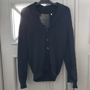 Marinblå stickad kofta från Stenströms - Marinblå stickad kofta från Stenströms i storlek S. Koftan har en bred krage, ribbade muddar och knäppning med fyra svarta knappar framtill. Materialet är grovstickat och ger en schysst struktur. Perfekt för dig som gillar klassisk stil med en modern touch.