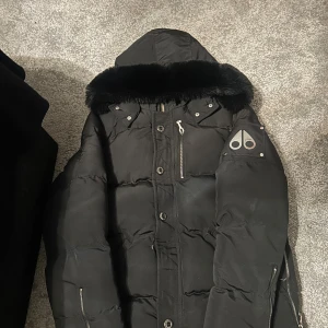 Svart dunjacka från Moose Knuckles - Svart dunjacka från Moose Knuckles med fluffig huva, silverfärgade dragkedjor och knappar. Jackan har logga på ärmen och flera fickor med dragkedja. Yttermaterial i bomull och nylon, fyllning av dun och fjäder. Perfekt för kalla dagar.