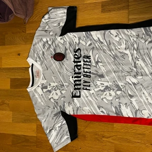 AC Milan vit och grå fotbollströja - Säljer en AC Milan fotbollströja i vit och grå med abstrakt mönster, svarta och röda detaljer på sidorna. Tröjan har korta ärmar, rund hals och klubbens emblem på bröstet. Materialet är lätt och andas, perfekt för träning eller match.