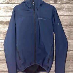 Blå vindjacka Peak Performance - Snygg marinblå vindjacka från Peak Performance med huva och hel dragkedja framtill. Jackan har två praktiska fickor med dragkedja och diskret logga på bröstet. Perfekt för blåsiga dagar och outdoor-aktiviteter.