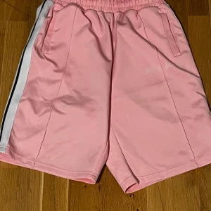 Rosa palm angels shorts - Säljer ett par ljusrosa sportiga shorts med vita och svarta ränder längs sidorna. De har elastisk midja och två fickor framtill. Perfekta för en avslappnad och trendig look under varma dagar.
