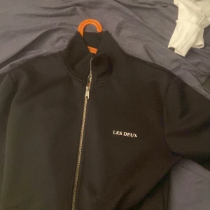 Svart zipjacka från Les Deux - Svart jacka från Les Deux med hög krage och hel dragkedja framtill. Stilren design med vit broderad logga på bröstet. Jackan har en clean look och är tillverkad i ett mjukt, slätt material som känns sportigt och modernt.