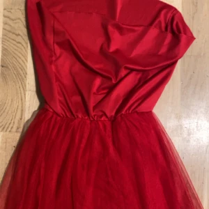 Röd axelbandslös balklänning - Snygg röd axelbandslös klänning med voluminös kjol i tyll och glansig överdel. Perfekt för dig som vill sticka ut med en klassisk och elegant look. Kjolen har ett lätt fluffigt fall och midjan är markerad för en smickrande siluett.